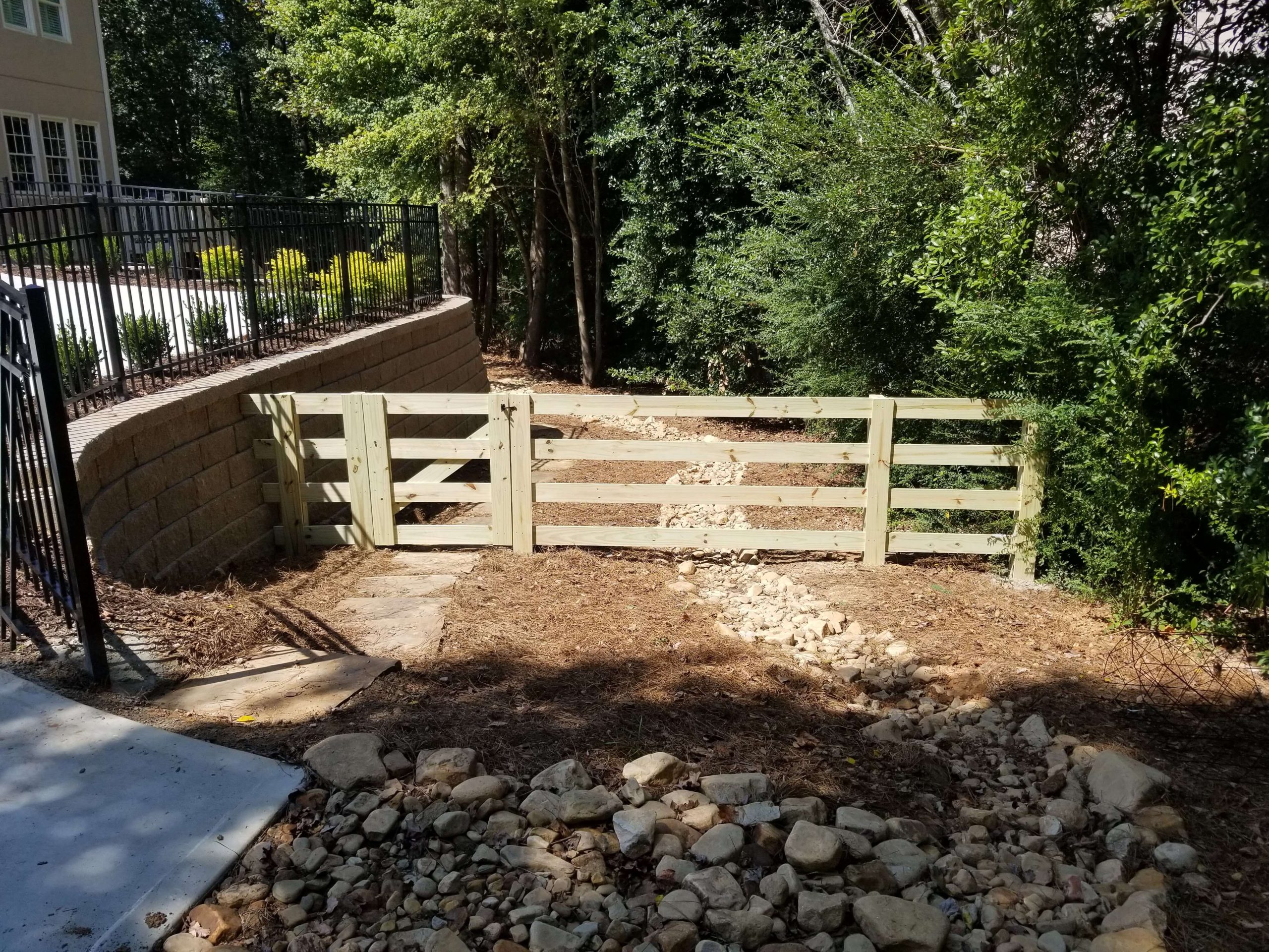 A wooden gate and fence border a rocky, wooded yard beside a house, with a black metal railing and concrete walkway on the left.