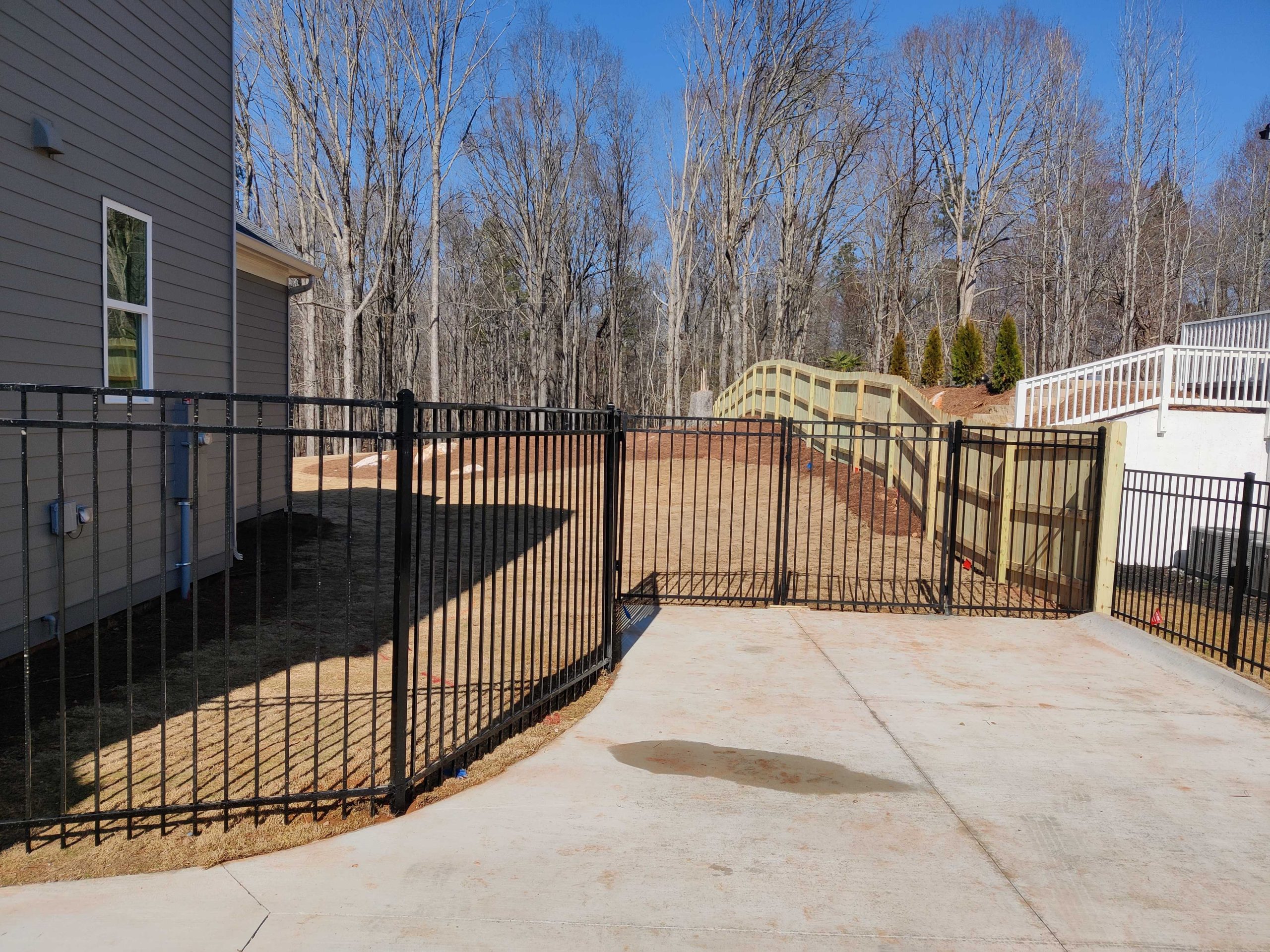 Black metal fencing encloses a concrete driveway beside a house, with a wooden footbridge and leafless trees in the backyard.